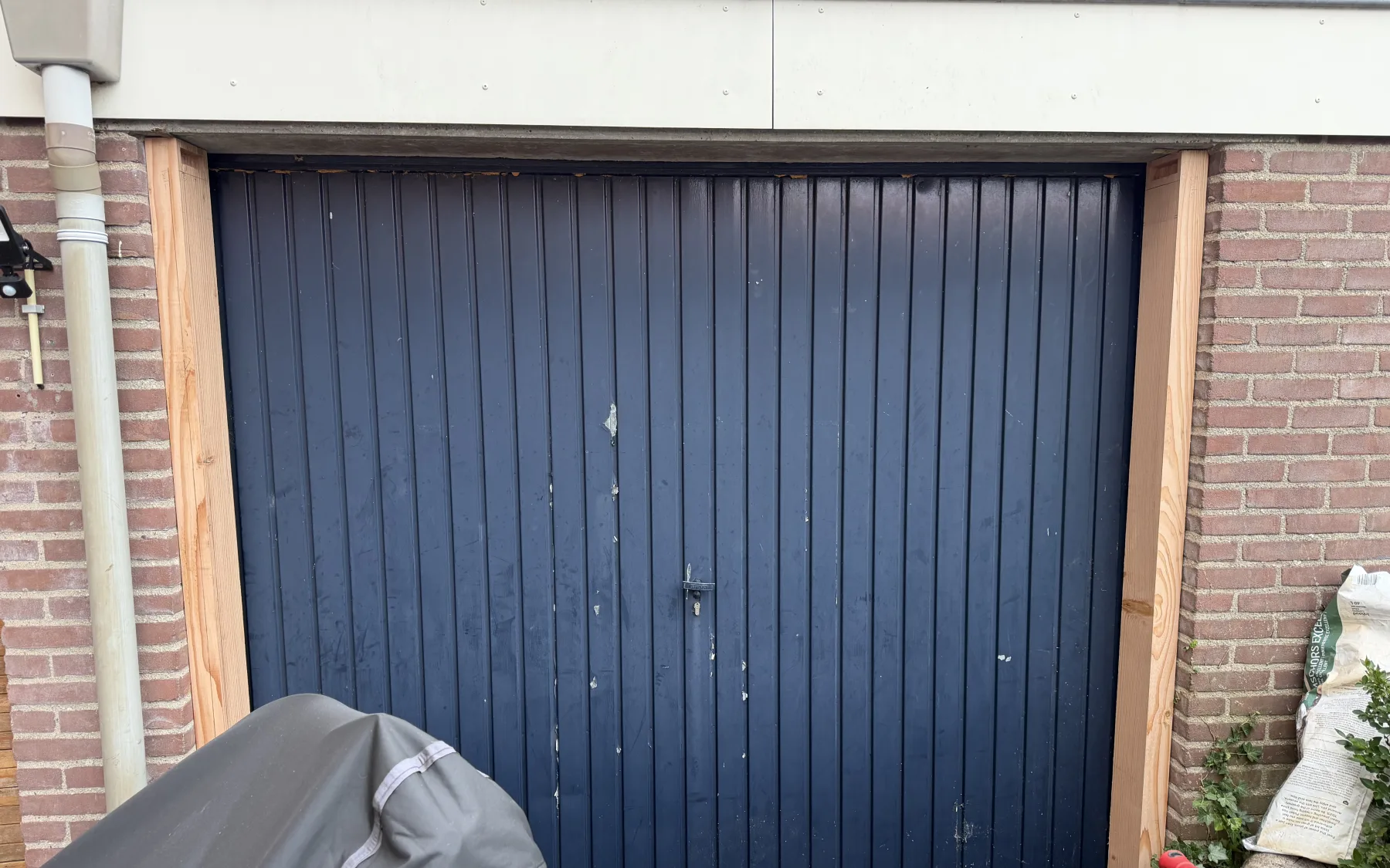 De oude blauw geschilderde stalen kanteldeur in de gemetselde garageopening, met de nieuwe Douglas kozijnstijlen er al naast